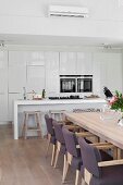 Dining area with upholstered chairs around wooden table in front of open-plan kitchen with hob on minimalist counter, fitted appliances and fitted cupboards with white fronts