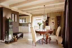 Dining area with period furniture below pendant lamp with large glass lampshade suspended from wood-beamed ceiling in elegant, open-plan interior; white country-house kitchen in background