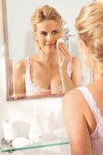 A blonde woman in front of a mirror cleaning her face with a cotton wool pad