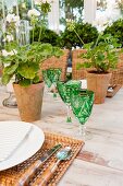 Ausschnitt aus gedecktem Tisch mit grünen Kristallgläsern, Bast-Sets, Terracotta-Töpfen mit Geranien