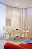 White Ghost Chairs at table in front of white curtain partition; orange sofa in foreground