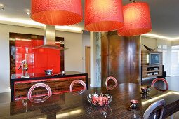 Orange lamps above metal chairs and large table with dark, exotic wood veneer; open-plan kitchen with red fitted cupboards in background