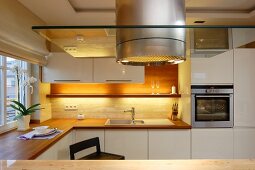 Stainless steel extractor hood with glass panel in modern kitchen with white cupboards and indirect lighting above kitchen counter