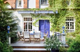 View from garden past table and chairs on terrace with lanterns on stone plinths to house with climber-covered façade and blue-painted wooden door