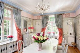 White table and chairs with carved backrests in dining room with decoratively painted walls and draped curtains on windows