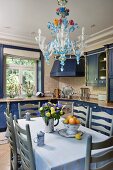 Esstisch mit hellgrauen Stühlen unter Kronleuchter aus farbigem Glas, in Küche mit blau lackierten Schränken