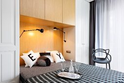 Double bed in niche of fitted wardrobes, black and white patterned bed linen and scatter cushions, carafe and glasses on tray