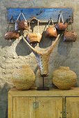Deko-Tierschädel zwischen Gefässen aus Geflecht auf hellem Holzschrank, darüber handgefertigte Glocken auf Wandhakenleiste an Lehmwand