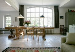 Long solid-wood table and simple chairs in front of arched lattice window in open-plan interior with rustic flair