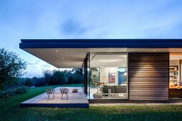 Architect-designed, classic modern house at twilight with roof projecting over terrace, glazed facade and illuminated interior