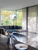 Retro metal teapot and place setting on table, corner sofa and arc lamp in background in front of glass wall with partially closed curtains