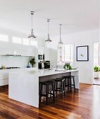 Stools at kitchen counter below retro pendant lamps in open-plan kitchen with white fronts and parquet floor