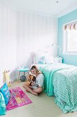 Girl playing guitar sitting next to bed with mint-green crocheted blanket in bedroom in pastel shades