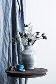 Trockenblumen in gestreifter Keramikvase auf rustikalem Bistrotisch vor Vorhang
