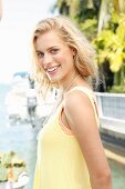 A blonde woman by the sea wearing a sleeveless, pastel yellow top