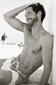 Lachender junger Mann in Badehose sitzt am Strand (s/w-Aufnahme)