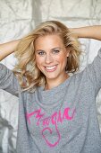 Blonde Frau in grauem Pulli mit pinkfarbener Schrift 'Magic' hält Arme hinter Kopf