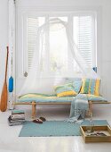A wooden lounger with striped upholstery and a mosquito net in front of a window in a light living room