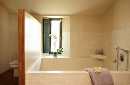 Large bathtub in corner of bathroom below window and toilet behind screen