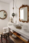 Rustic wooden stool in front of white washstand with twin basins below vintage mirror with ornate gilt frame