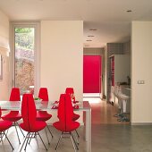 Esstisch und rote, gepolsterte Designerstühle auf Estrichboden, Blick durch raumhohen Durchgang in Küche mit Barhockern vor Theke