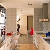 White designer kitchen with pale grey wall tiles, open doorway with view of woman walking down staircase in living room