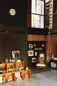 Double-height loft apartment with collection of spirits in crate-style shelving; gymnastic rings hanging form iron mezzanine balustrade and antique armchairs in corner