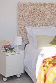 Teilweise sichtbares Bett vor Raumteiler mit Wandbehang, daneben mobiler Nachttisch auf Rädern und kleine, weiße Tischleuchte