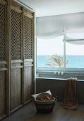 Fitted cupboards with rustic wooden doors with mesh panels next to bathtub below window with sea view