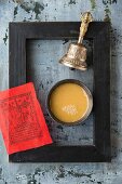 A bowl of butter tea, a red prayer flag and a bell inside a black picture frame (Tibet)