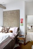 Antique bedside table next to bed against tall headboard panel upholstered in pale grey