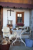 Weißer Vintage Gartentisch und Gartenstuhl mit Schaffell vor Bauernhaus, auf Tisch Blumentöpfe mit Heidekraut und Tannenzapfen