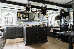 Spacious kitchen with black-painted, traditional-style base units and island counter below cooking utensils hung from ceiling