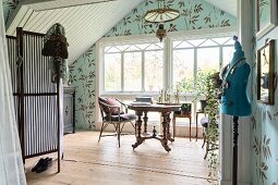 Nostalgisch gestaltetes Giebelzimmer mit Schneiderpuppe, im Hintergrund antiker Tisch vor Sprossenfenster, an Wand Tapete mit Blättermotiv