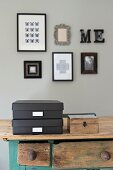 Black storage boxes with labels and wooden box on table below framed pictures on grey wall