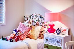Soft toys and scatter cushions on bed with headboard upholstered in floral fabric and pink table lamp on bedside cabinet