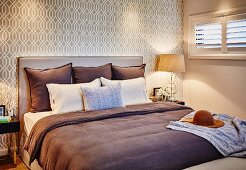 Doppelbett mit brauner Tagesdecke, Kissensammlung und gepolstertem Kopfteil vor beigefarbener Retrotapete