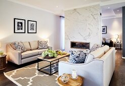 Elegant sofa set with pale covers and modern coffee table on rug with large ornamental pattern in open-plan interior with fireplace in partition wall