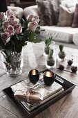 Small gift and tealight holders on stacked books on tray in front of glass vase of roses on coffee table