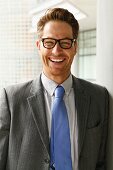 A man with a wide grin wearing glasses, a grey blazer and a tie