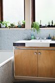 Retro-Waschtisch aus hellem Holz mit eingebautem Waschbecken vor Fenster, helle Mosaikfliesen an Wand und Badewanne