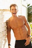 A young, topless, tattooed man standing against a wall laughing
