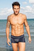 A young, tattooed man by the sea wearing bathing trunks