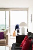 Black sofa with scatter cushions in shades of red, white table lamp on side table, open sliding terrace doors showing man and dog in background