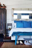 Vintage mirror on white wooden wall above bed with blue and white patterned scatter cushions and blue bench at foot