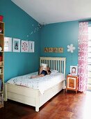 Child on white bed in corner of blue-painted room with framed pictures and letters on wooden boards on walls