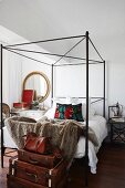 Stack of vintage suitcases at foot of black metal four-poster bed in modern bedroom