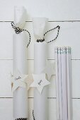 Weiß verpackte Geschenkrollen mit beschrifteten Geschenkanhängern und Bleistifte auf weißem Holzuntergrund