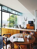 Round, wooden, antique table and armchairs in front of free-standing, wooden sideboard; hunting trophies on wall of lounge area in background