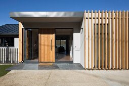 Roofed porch of contemporary house with central, plain wood front door, glass walls and wooden structure in front of facade